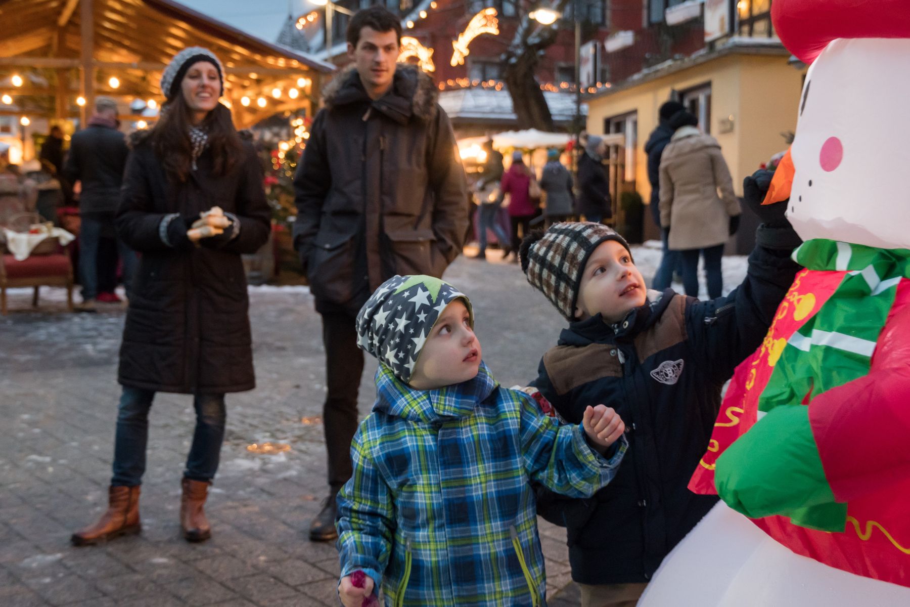 Zwei Jungen in Winterkleidung betrachten aufgeregt einen aufblasbaren Schneemann auf einem Weihnachtsmarkt im Freien, während ein Mann und eine Frau hinter ihnen stehen, mit festlichen Lichtern und Menschen im Hintergrund.